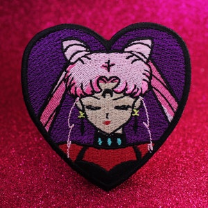 Puede incluir: Un parche bordado en forma de corazón morado con un personaje de anime de pelo rosa, una corona y una gargantilla negra. El personaje tiene los ojos cerrados y una boca roja.