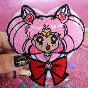 K&ouml;nnte beinhalten: Rosa und roter gestickter B&uuml;gelaufkleber mit einer Comicfigur mit herzf&ouml;rmigem Kopf, die eine Matrosenuniform und eine rote Schleife tr&auml;gt. Die Figur hat rosa Haare, rote Augen und einen Stern auf der Stirn.