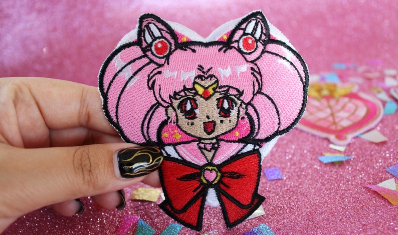 K&ouml;nnte beinhalten: Rosa und roter gestickter B&uuml;gelaufkleber mit einer Comicfigur mit herzf&ouml;rmigem Kopf, die eine Matrosenuniform und eine rote Schleife tr&auml;gt. Die Figur hat rosa Haare, rote Augen und einen Stern auf der Stirn.