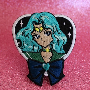May include: An embroidered iron-on patch featuring a heart-shaped frame with a cartoon image of a Sailor Moon character with blue hair and a green and blue outfit. The character has a yellow star on her forehead and a blue bow with a green gem in the center.