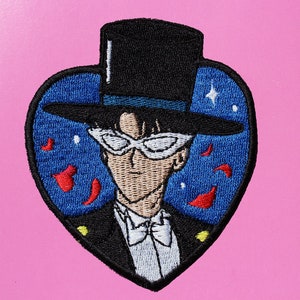 Może przedstawiać: Naszywka haftowana do prasowania z postacią z anime Sailor Moon. Postać ma na sobie czarny cylinder, biały smoking i białe okulary. Tło jest niebieskie z czerwonymi płatkami róż i białymi gwiazdami.