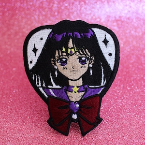 Può includere: Toppa ricamata raffigurante un personaggio in stile anime con capelli neri, occhi viola e un fiocco rosso scuro. La toppa ha un bordo nero con dettagli bianchi, su uno sfondo rosa glitterato.