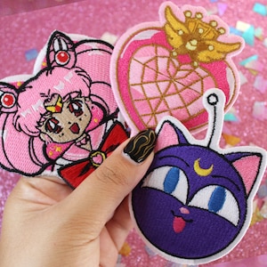 May include: Three embroidered iron-on patches featuring characters from the anime Sailor Moon. The patches are pink, purple, and gold. The first patch is a pink Sailor Moon with a red bow and a gold crown. The second patch is a pink heart with a gold crown and a web-like design. The third patch is a purple cat with a crescent moon on its forehead.