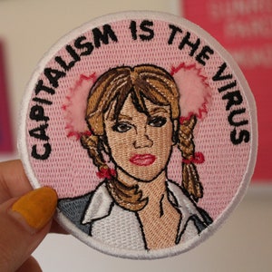 May include: A pink and white embroidered patch featuring a cartoon image of a girl with pigtails and the text "Capitalism is the Virus".