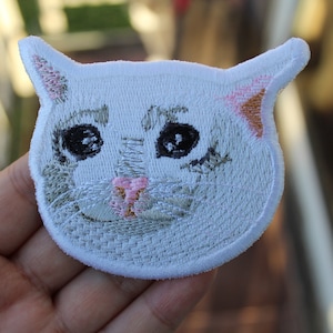 Puede incluir: Parche de gato blanco bordado con ojos negros, nariz rosa y una oreja marrón claro. El parche está en una mano.