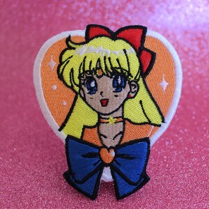 Puede incluir: Un parche bordado para planchar con un personaje de anime de dibujos animados con cabello amarillo, un lazo rojo y un lazo azul. El personaje lleva un atuendo naranja y amarillo y está rodeado de un corazón blanco con fondo naranja.