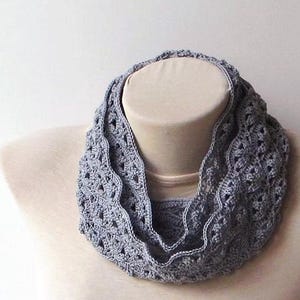 May include: A gray crocheted infinity scarf displayed on a mannequin. The scarf has a delicate, openwork design with a scalloped edge. The soft, muted gray color and intricate pattern suggest a handmade item, perfect for adding a touch of elegance to any outfit.