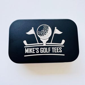 May include: A rectangular black metal tin with rounded corners featuring a white graphic of a golf ball on a tee flanked by two golf flags and two golf clubs. The text MIKE'S GOLF TEES is printed in white capital letters below the graphic.