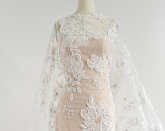 May include: A sheer white fabric with intricate floral embroidery, draped over a mannequin. The design features large, three-dimensional white flowers and delicate embellishments. The Couture Lace logo is visible.