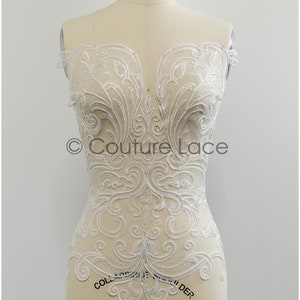 Puede incluir: Aplique de encaje blanco con un diseño floral. El encaje está adornado con cuentas y tiene un patrón delicado e intrincado. El aplique se muestra en un maniquí blanco.