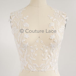 May include: White floral lace applique with a delicate floral pattern. The lace is shown on a dress form and is perfect for adding a touch of elegance to any garment.