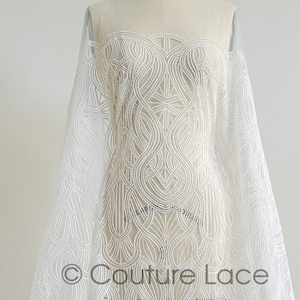 Puede incluir: Tela de encaje blanca con un intrincado patrón fluido drapeada sobre un maniquí. El delicado diseño presenta un motivo arremolinado repetitivo. La tela es transparente, permitiendo el paso de la luz, e ideal para novias o ropa formal.