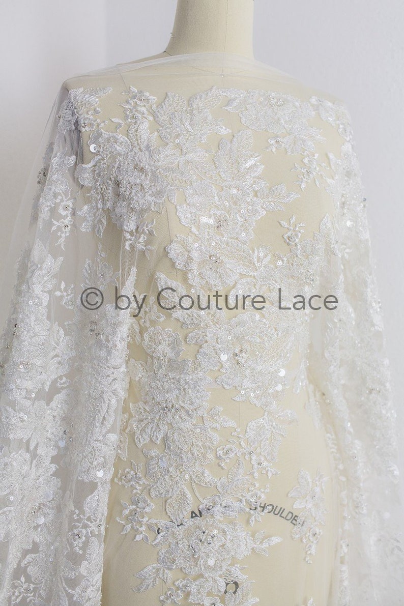 May include: White floral lace fabric with sequins and beads. The lace has a delicate floral pattern and is perfect for wedding dresses, bridal gowns, and other special occasion garments.