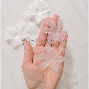May include: A hand holding two white, delicate, paper-like flower shapes. The flower shapes are translucent and have a slightly crinkled texture. The background is white.