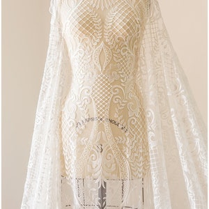 K&ouml;nnte beinhalten: Wei&szlig;er Spitzenstoff mit einem zarten Blumenmuster. Die Spitze befindet sich auf einer Schaufensterpuppe und hat einen gewellten Rand.