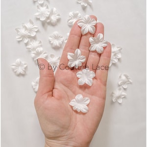 Könnte beinhalten: Eine Hand hält eine Sammlung weißer Satinblütenverzierungen. Die Blüten sind auf einer weißen Oberfläche verstreut. © by Couture Lace