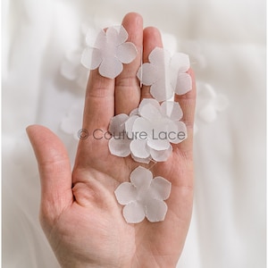 May include: A hand holding several white fabric flower embellishments. The flowers are delicate and have a slightly ruffled texture.