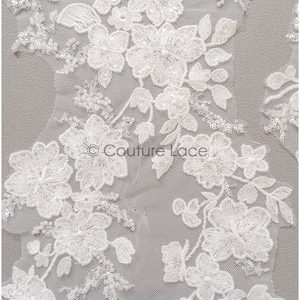 Op de afbeelding: Close-up van witte bloemenkantstof. Het kant heeft gedetailleerde geborduurde bloemen, bladeren en kralenaccenten. Het delicate ontwerp staat tegen een transparante achtergrond, waardoor een textuur en elegante uitstraling ontstaat. Het Couture Lace-logo is zichtbaar.
