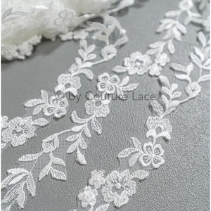 May include: White floral lace trim with a delicate design of small flowers and leaves. The lace is on a gray background.