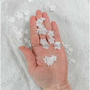 May include: A hand holding a collection of small, white, fabric flower embellishments. The flowers are delicate and have a slightly ruffled texture.