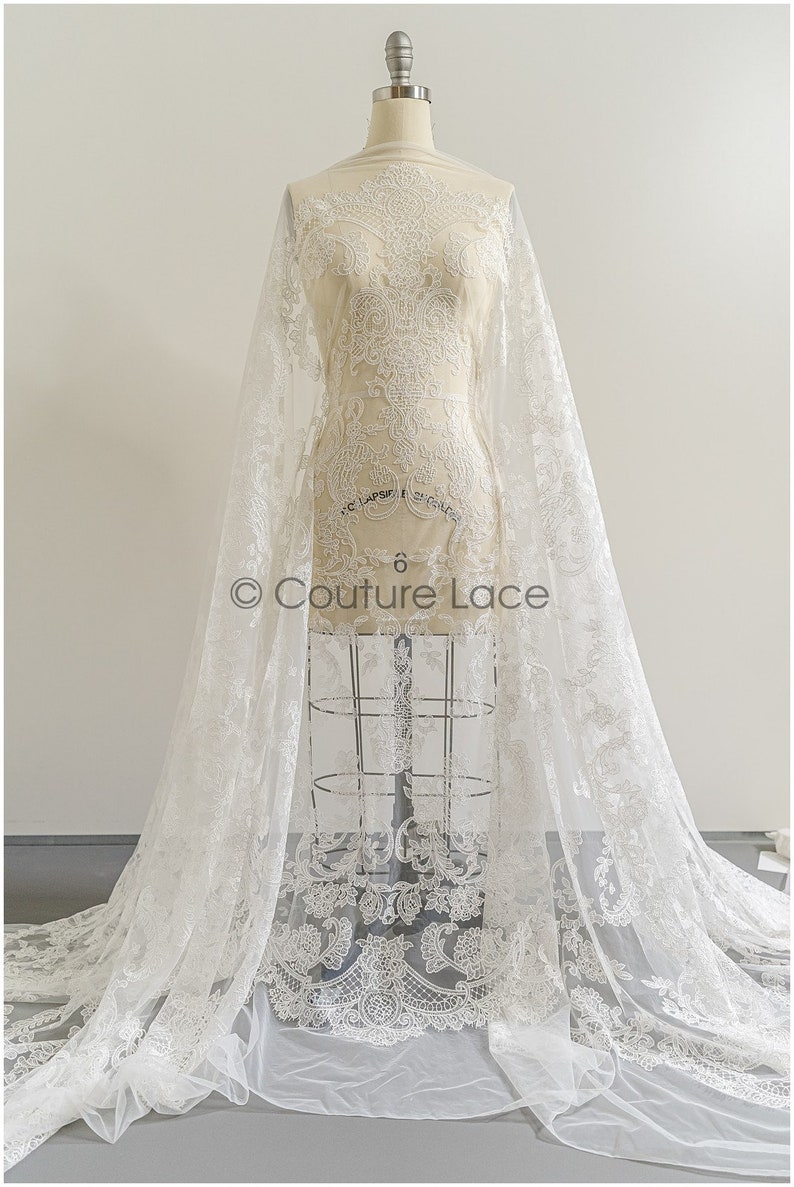 May include: White lace fabric with a delicate floral pattern. The lace is shown draped over a dress form. The fabric is sheer and has a soft, flowing texture. The text "Couture Lace" is visible on the fabric.