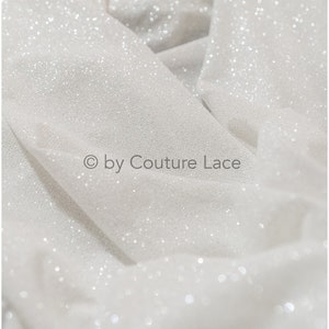 May include: White fabric with a sparkly, glittery texture. The fabric is folded and has a subtle sheen. The text "&copy; by Couture Lace" is visible in the lower center of the image.
