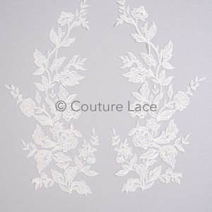May include: Two white lace appliques with floral designs. The appliques are shaped like a pair of branches with leaves and flowers.