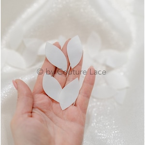 May include: Close-up of several white, petal-shaped fabric pieces held in a hand. The petals are smooth and opaque, with a slight curve. The background is a white, textured fabric. The text "© by Couture Lace" is visible.