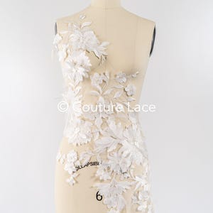Pode incluir: Aplique de renda floral branca com flores e folhas delicadas. A renda está sobre um fundo de malha transparente. © Couture Lace