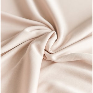 May include: A close-up of a smooth, light beige fabric with soft folds and wrinkles. The fabric has a subtle sheen and a luxurious feel.