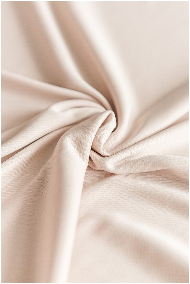 May include: A close-up of a smooth, light beige fabric with soft folds and wrinkles. The fabric has a subtle sheen and a luxurious feel.