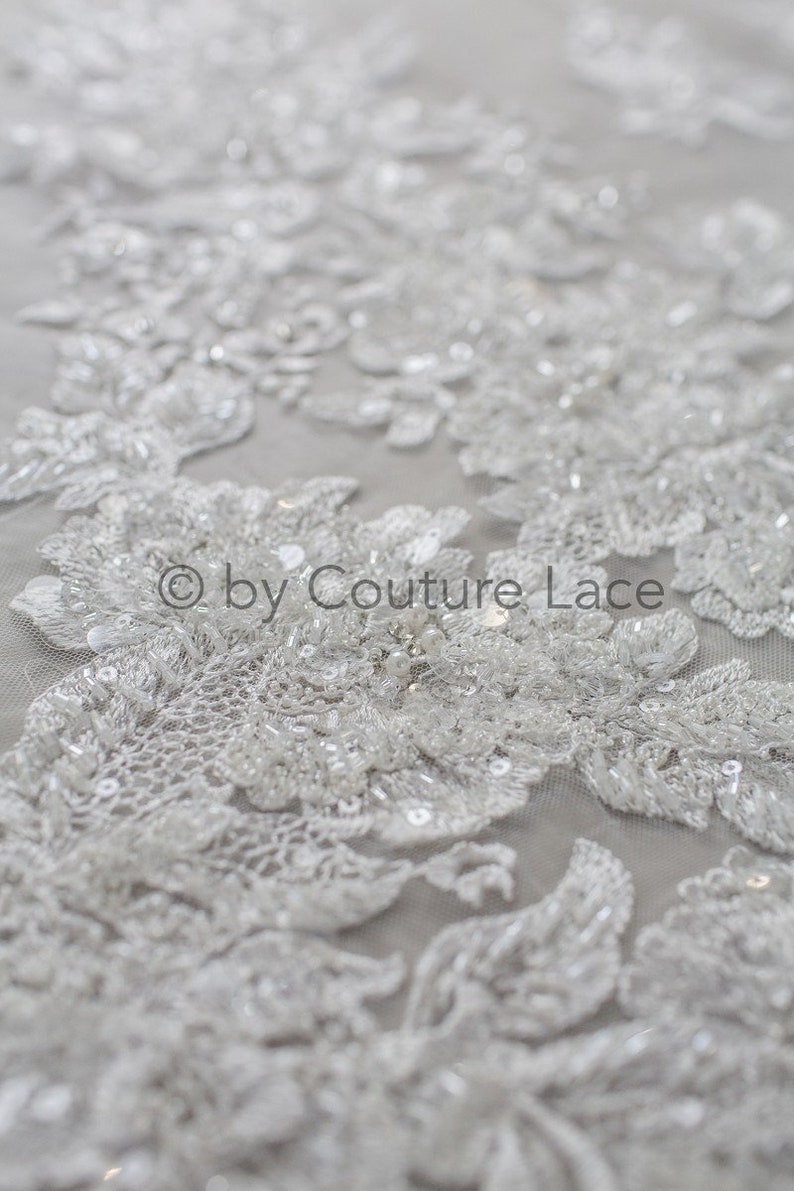 May include: White beaded lace fabric with a floral design. The lace is delicate and intricate, with small beads and sequins scattered throughout. The fabric is perfect for wedding dresses, bridal accessories, and other special occasion garments.