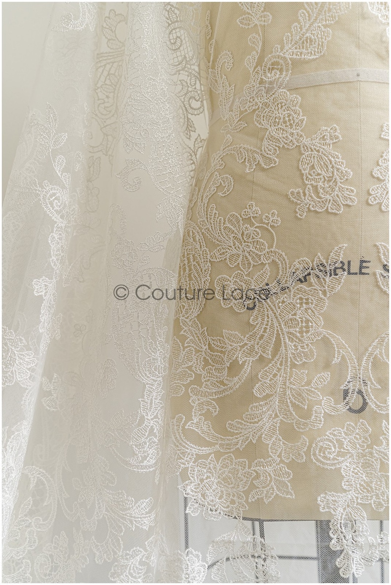 May include: White lace fabric with a floral pattern. The lace is delicate and intricate, with a soft, flowing texture. The fabric is shown draped over a dress form.