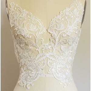 May include: White floral lace applique with a V-neck design. The delicate lace features intricate patterns of flowers and leaves, perfect for bridal or formal wear. The applique is displayed on a mannequin.