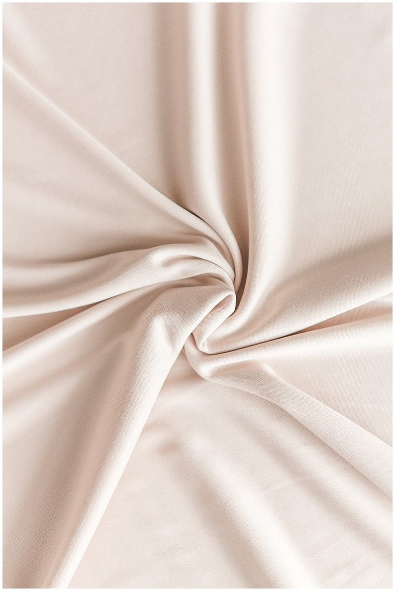 May include: A close-up of a smooth, pale pink fabric with soft folds and wrinkles. The fabric is draped and gathered in a swirling pattern, creating a luxurious and elegant texture.