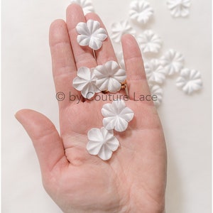 May include: A hand holding a collection of small, white, fabric flower embellishments. The flowers are delicate and have a ruffled texture.  The flowers are scattered on a white surface.