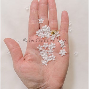 Könnte beinhalten: Eine Hand hält einen Haufen kleiner weißer Stoffblumen. Die Blumen sind zart und haben eine leicht gekräuselte Textur. Der Text "© by Couture Lace" ist im Bild sichtbar.