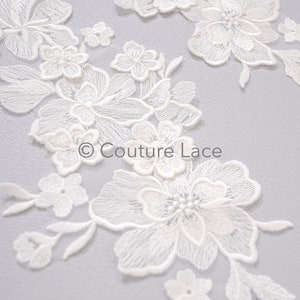 Pode incluir: Aplique de renda floral branca com detalhes delicados de bordado. A renda apresenta uma variedade de formas e tamanhos de flores, com uma flor maior no centro. A renda é perfeita para adicionar um toque de elegância a qualquer projeto.