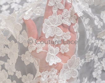 May include: White floral lace fabric with a delicate, embroidered design. The lace is sheer and has a soft, flowing texture.
