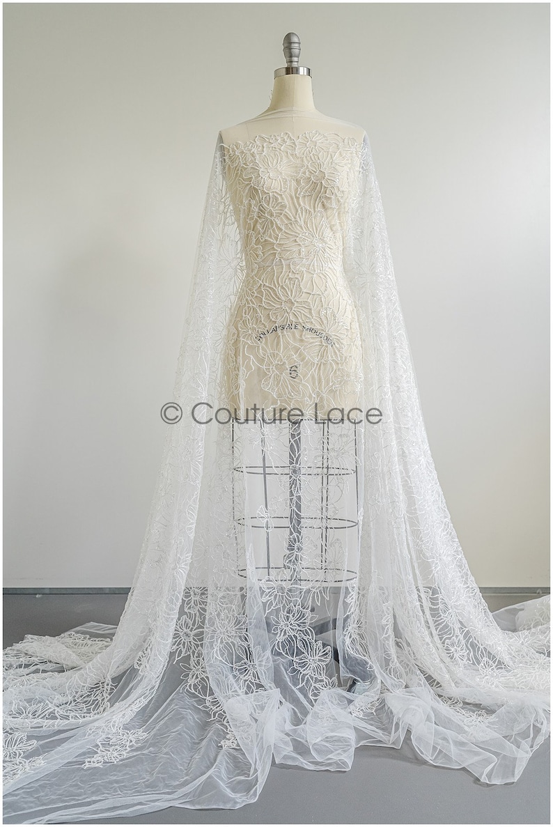 Pu&ograve; includere: Tessuto di pizzo floreale bianco con un motivo delicato. Il pizzo &egrave; su un manichino bianco e drappeggiato sul pavimento.