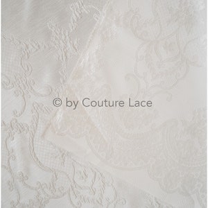May include: White fabric with a raised, embossed pattern of a floral design. The fabric is folded in half, and the text "&copy; by Couture Lace" is visible in the center of the image.