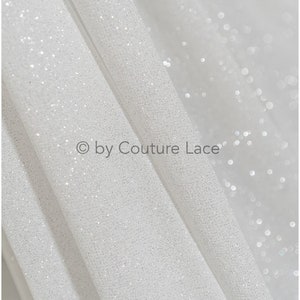 May include: White glitter fabric with a subtle shimmer. The fabric is folded in a way that shows the texture of the glitter. The text "&copy; by Couture Lace" is visible in the image.