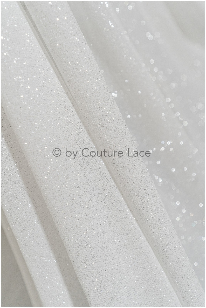 May include: White glitter fabric with a subtle shimmer. The fabric is folded in a way that shows the texture of the glitter. The text "&copy; by Couture Lace" is visible in the image.