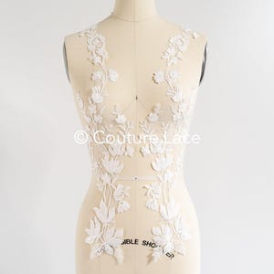 May include: White floral lace applique for a wedding dress or other garment. The lace is delicate and features a floral design with small pearls. The lace is shown on a dress form.