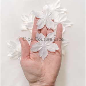 May include: A hand holding five white fabric flower embellishments. The flowers are delicate and have a slightly ruffled texture.