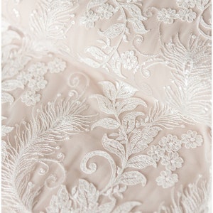 May include: Close-up of delicate white lace fabric with a floral pattern. The lace features intricate designs of leaves, flowers, and swirls.