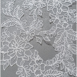 May include: Close-up of white floral lace fabric with intricate designs. The lace features detailed floral and leaf patterns, with delicate sequin accents. The design is set against a light gray background, highlighting the lace's texture and detail.