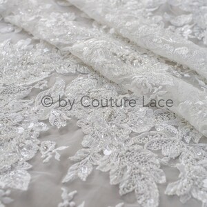 May include: White beaded lace fabric with a floral pattern. The lace is delicate and intricate, with small beads and sequins scattered throughout. The fabric is sheer and lightweight, making it ideal for special occasion garments.