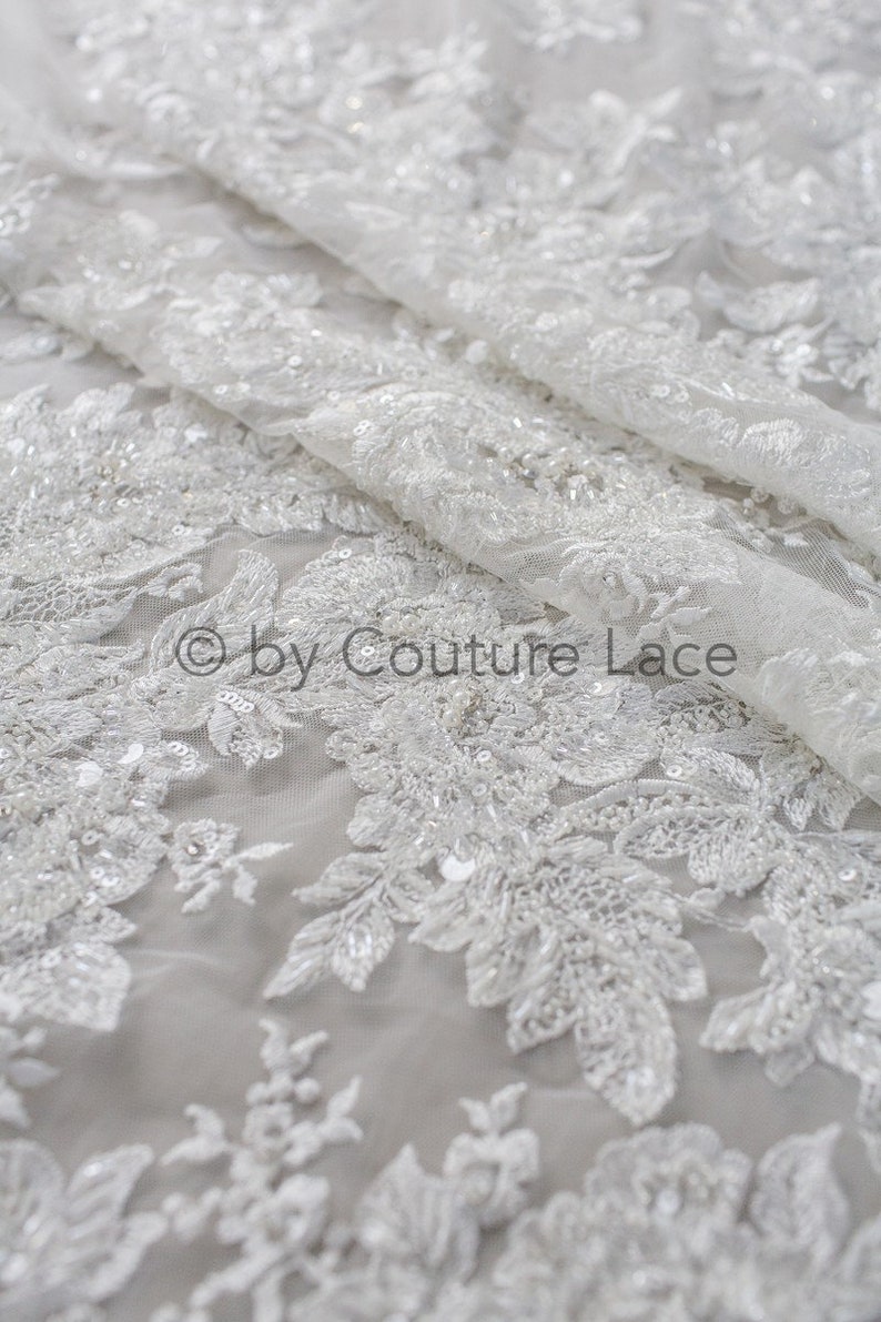 May include: White beaded lace fabric with a floral pattern. The lace is delicate and intricate, with small beads and sequins scattered throughout. The fabric is sheer and lightweight, making it ideal for special occasion garments.