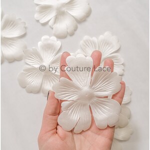 May include: A hand holding a white satin flower with 7 petals. The flower is surrounded by other white satin flowers on a white background. The text "© by Couture Lace" is visible in the image.